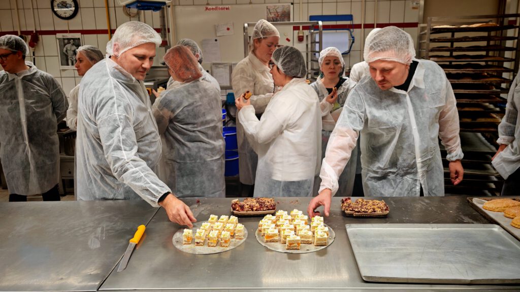Teilnehmende der Backhaus-Besichtigung 2025 greifen in Hygienekleidung zu kleinen Kuchenstücken in der Konditorei.