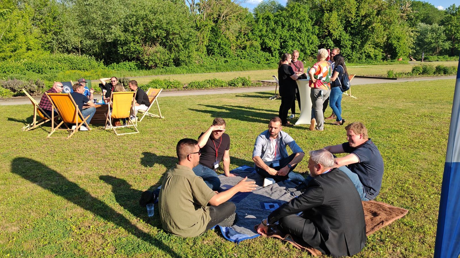 Gespräche auf der Wiese bei der WJ Business Lounge Teilnehmende der WJ Business Lounge sitzen auf einer Picknickdecke im Gespräch, dahinter weitere Gruppen an Liegestühlen und Stehtischen.